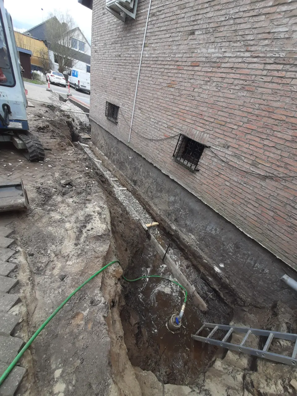 Freigelegter Arbeitsraum entlang einer Hauswand mit Minibagger und anstehendem Wasser